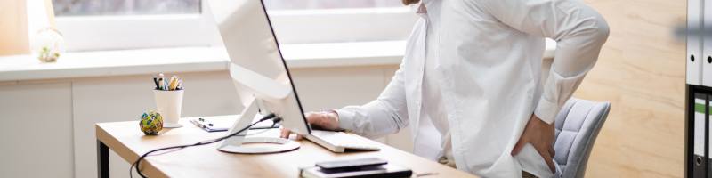 Fysiotherapie bij Houdingsproblemen Persoon die aan het werk is achter een bureau en de onderrug vasthoudt als teken van rugklachten